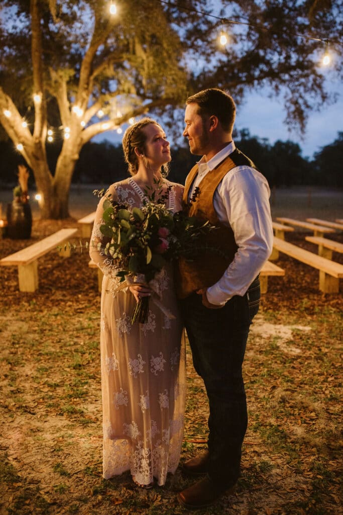 bride and groom standing outside by benches under large oak tree with market lights
